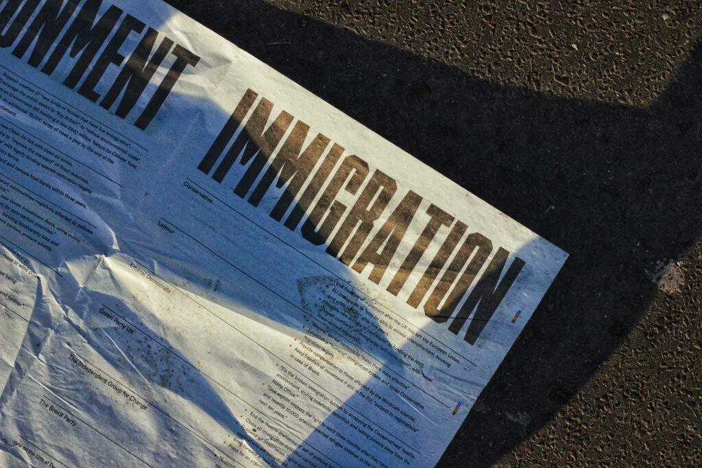 A crumpled newspaper page lying on asphalt, with the bold headline “IMMIGRATION” in large dark letters partially lit by sunlight and showing faint shadows across the printed text.