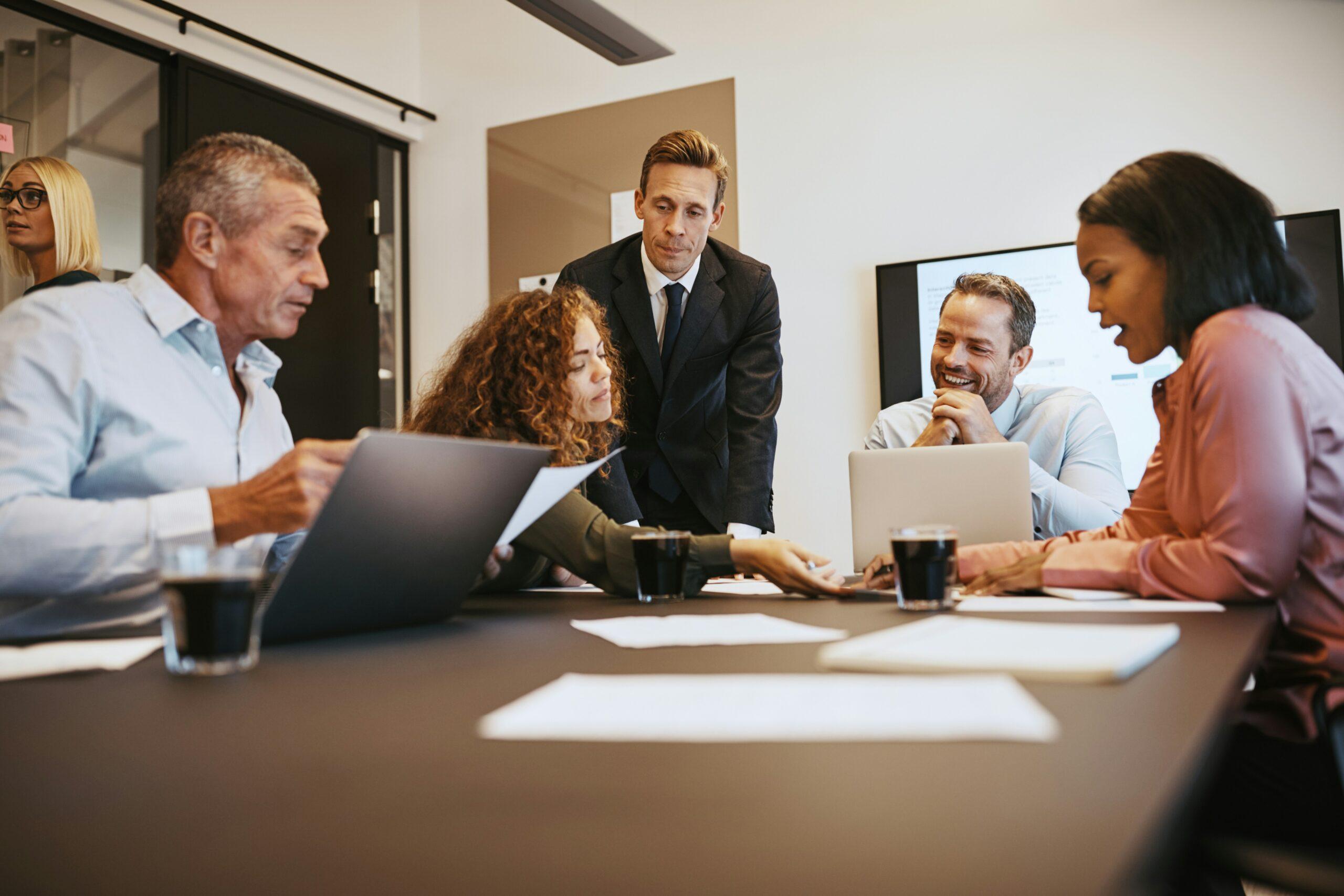 A group of professionals gathered around a conference table during a meeting, reviewing documents and laptops while discussing ideas in a modern office setting.