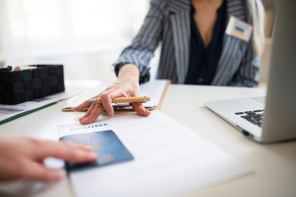 A close-up view of a person sitting at a desk during an administrative or hiring process, with one hand holding pens over a clipboard and the other person handing over a passport on top of a printed résumé, while a laptop and office supplies sit nearby.