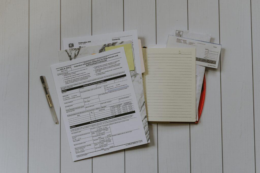 A collection of tax-related documents, forms, envelopes, and a lined notebook arranged on a light wooden surface, with a pen placed beside the paperwork.