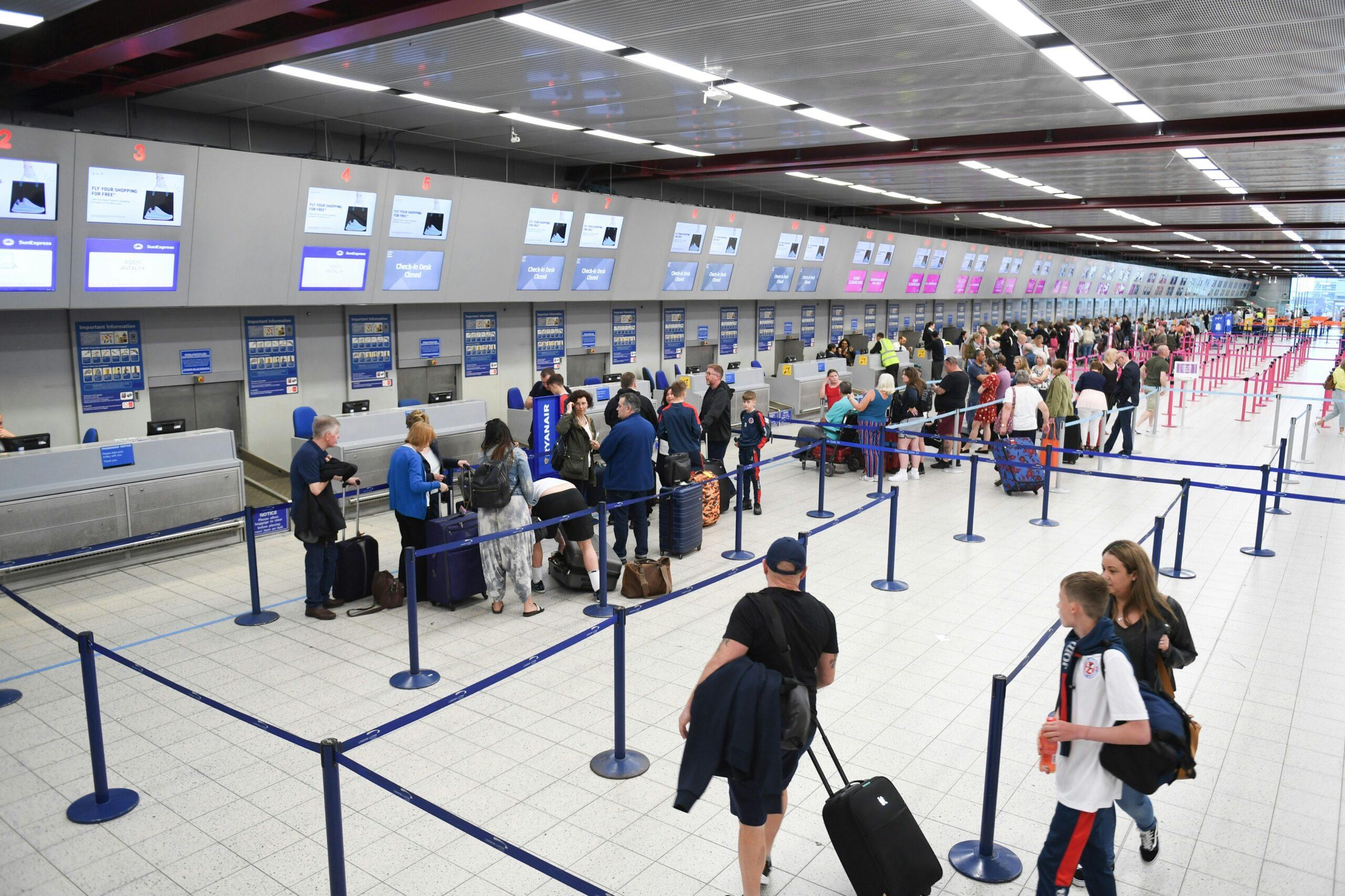 A wide view of a busy airport check-in area with long lines of travelers waiting with luggage, guided by blue queue barriers, while overhead screens display flight and counter information across the entire terminal.