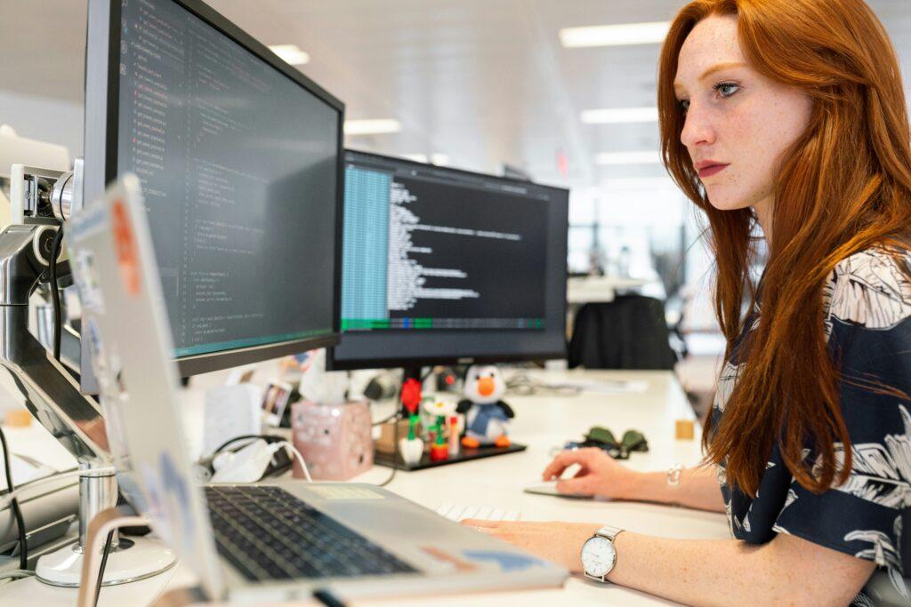 A focused software developer working at a desk with multiple monitors displaying code, using a laptop and mouse in a modern, well-lit office environment.