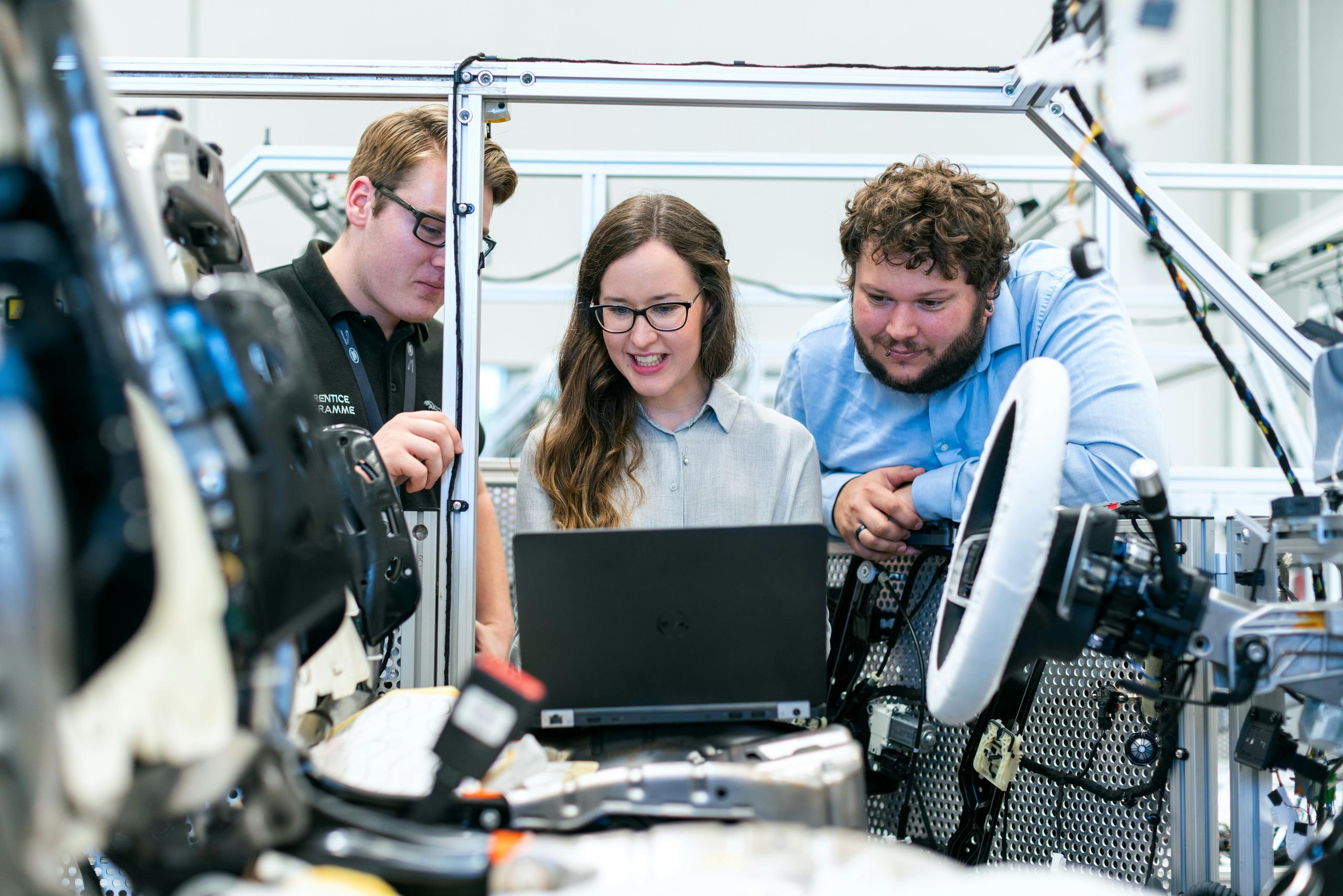 A group of three engineers gathered around a laptop inside an automotive testing frame, reviewing data while surrounded by wiring, mechanical components, and a mounted steering wheel.