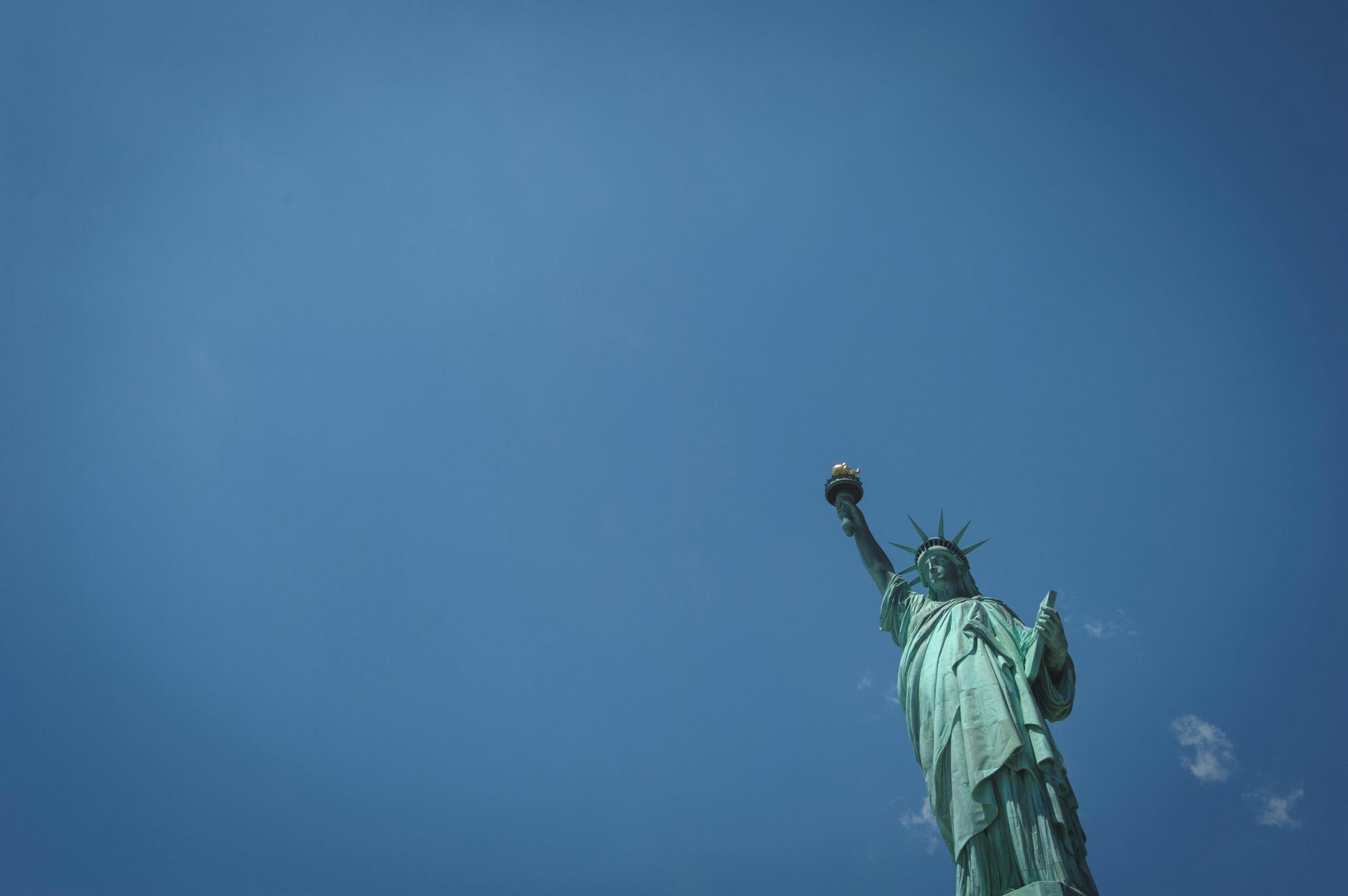 The Statue of Liberty rising against a clear blue sky, with its torch held high and only the upper portion of the monument visible in the frame.