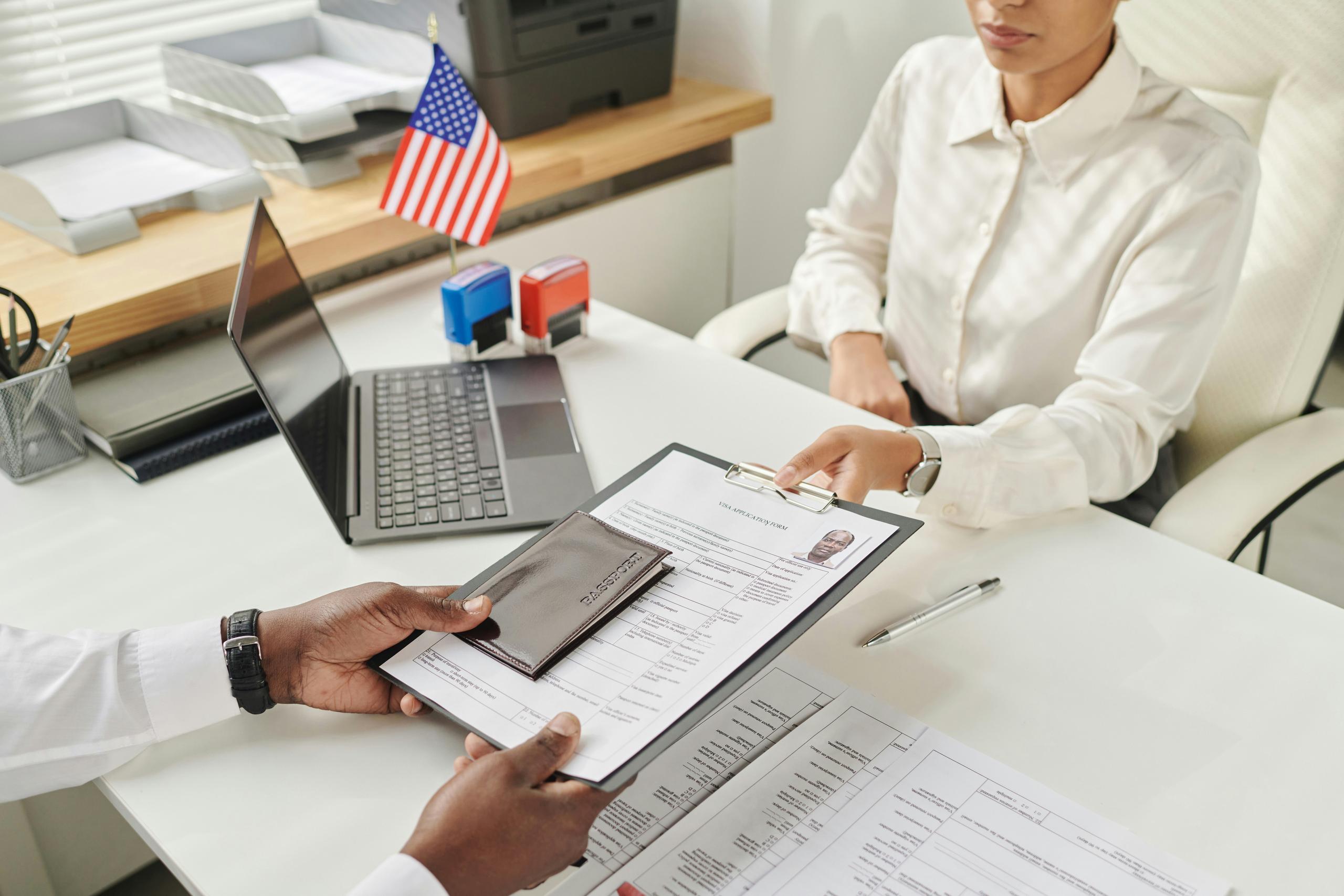 A person seated at a white office desk hands a clipboard holding a completed form and a passport to another individual across the desk, with a small U.S. flag, an open laptop, ink stamps, stacked document trays, and additional paperwork visible in the background, suggesting an official administrative or immigration-related process taking place in a professional office setting.