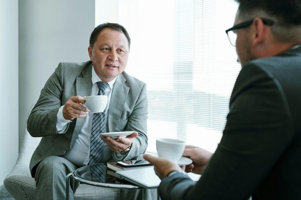 Two men in business suits sitting at a table, each with a coffee cup, engaged in conversation.